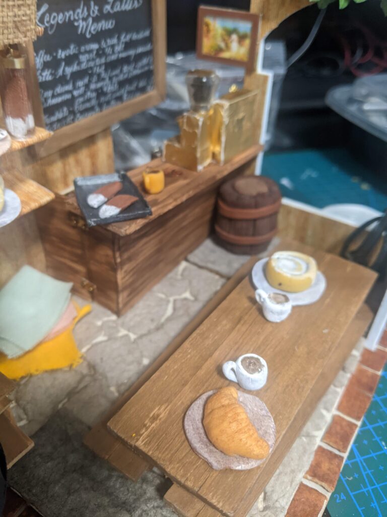 View of the front of Viv's coffee shop where we see a croissant, huge cinnamon roll and a couple of coffees on the trestle table. On the counter, we spot a couple of huge biscotti chilling on the pan and an iced coffee, paid for with two bits. 