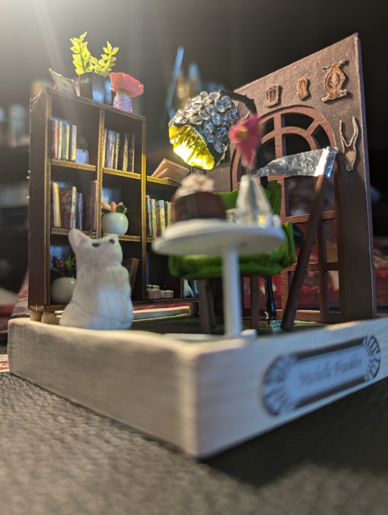 The miniature is lit with a single floor light, and none of the books are glued into the book cases, so it became a bit of an anti-stress exercise when I handed the miniature over to the author, along with a pair of tweezers. Unfortunately, the chair is glued down, so it makes the back shelf difficult to update.